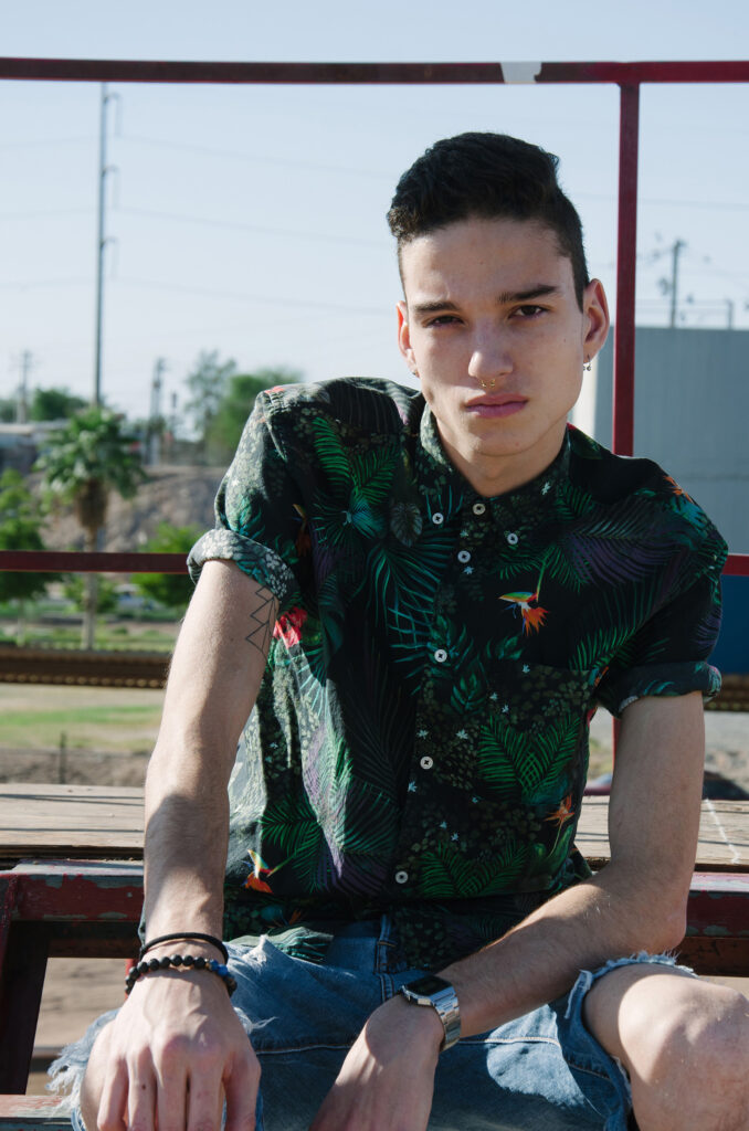Victor Zepeda a model sits on a rusty structure wearing a tropical-patterned shirt and distressed jeans with a tattoo on his arm and a watch on his wrist.
